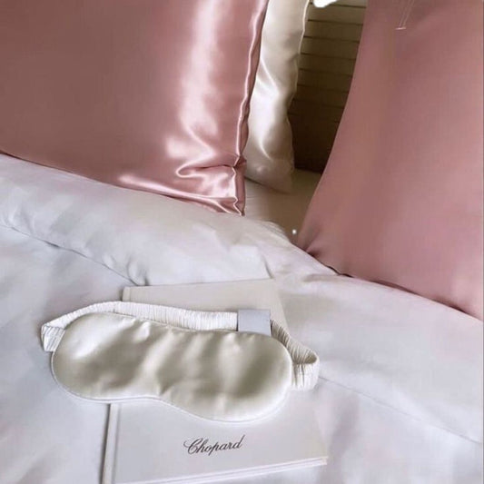 fundas de almohada de seda de color rosa y beige en una cama de matrimonio, con un libro y un antifaz de seda blanco