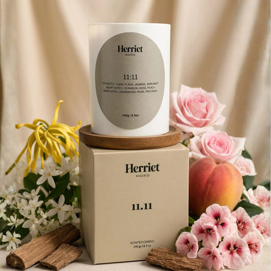 A white scented candle called 11.11 sitting on top of a green candle box, from Herriet, styled with pink roses, a peach, white and yellow flowers, cinnamon sticks, and a draped cream fabric background.