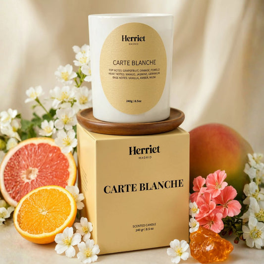 A scented candle with a yellow oval label sitting on top of a yellow Herriet scented candle box. The scene is styled with citrus slices, white flowers, pink blossoms, and a peach against a soft cream background.