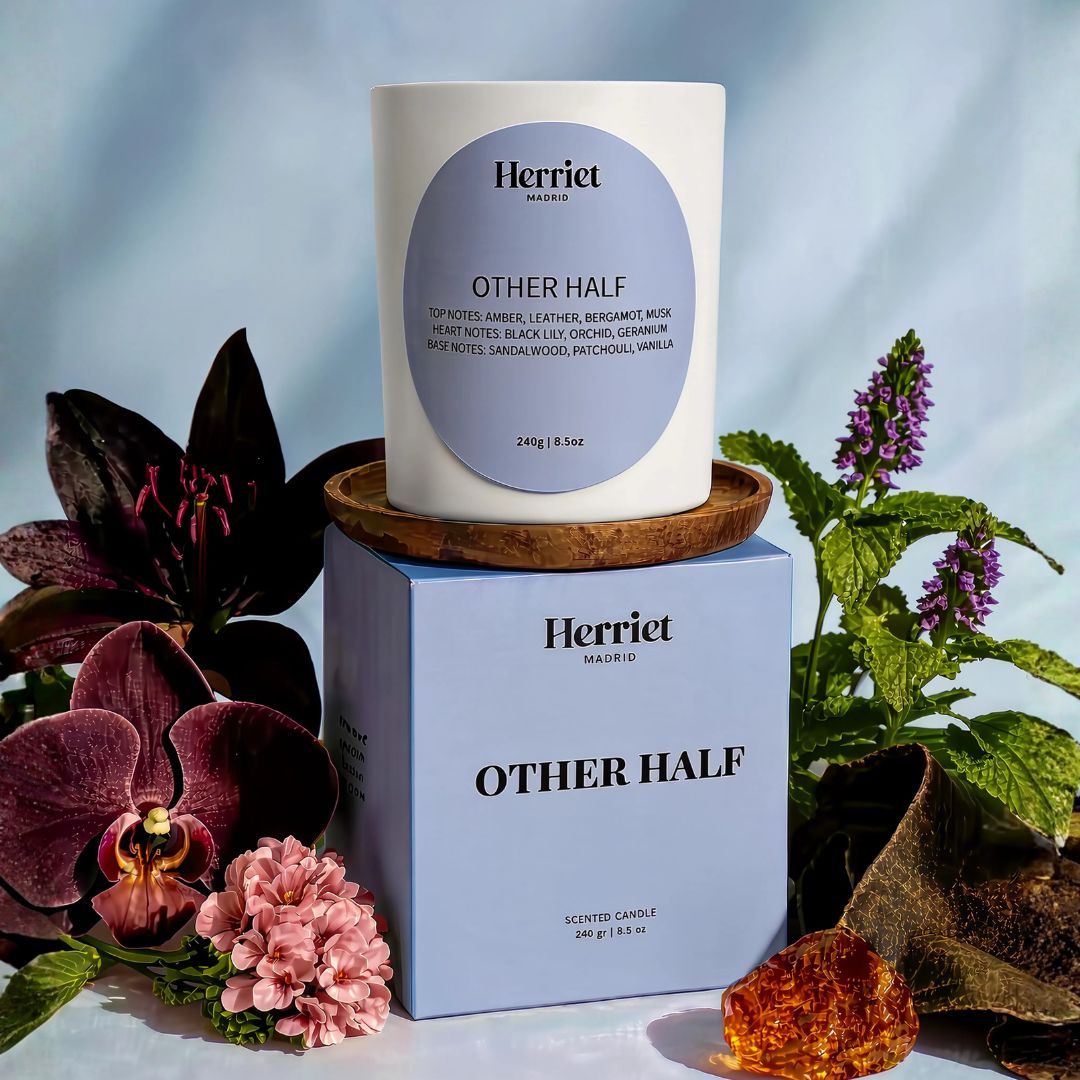 A white scented candle labeled 'Other Half' form Herriet, styled with flowers and greenery against a soft blue backdrop.