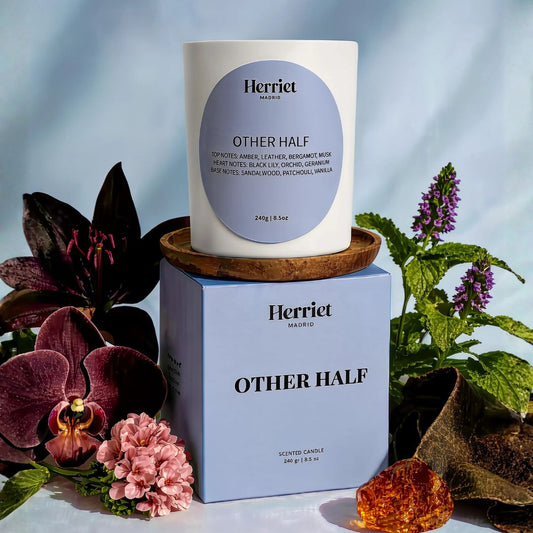 A white scented candle labeled 'Other Half' form Herriet, styled with flowers and greenery against a soft blue backdrop.