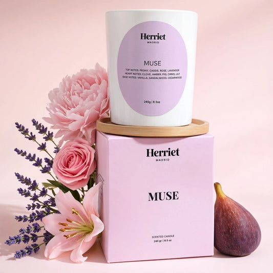 A white scented candle from Herriet called MUSE sitting on top of a pale pink candle box, styled with pink flowers and a fig on a soft pink background.