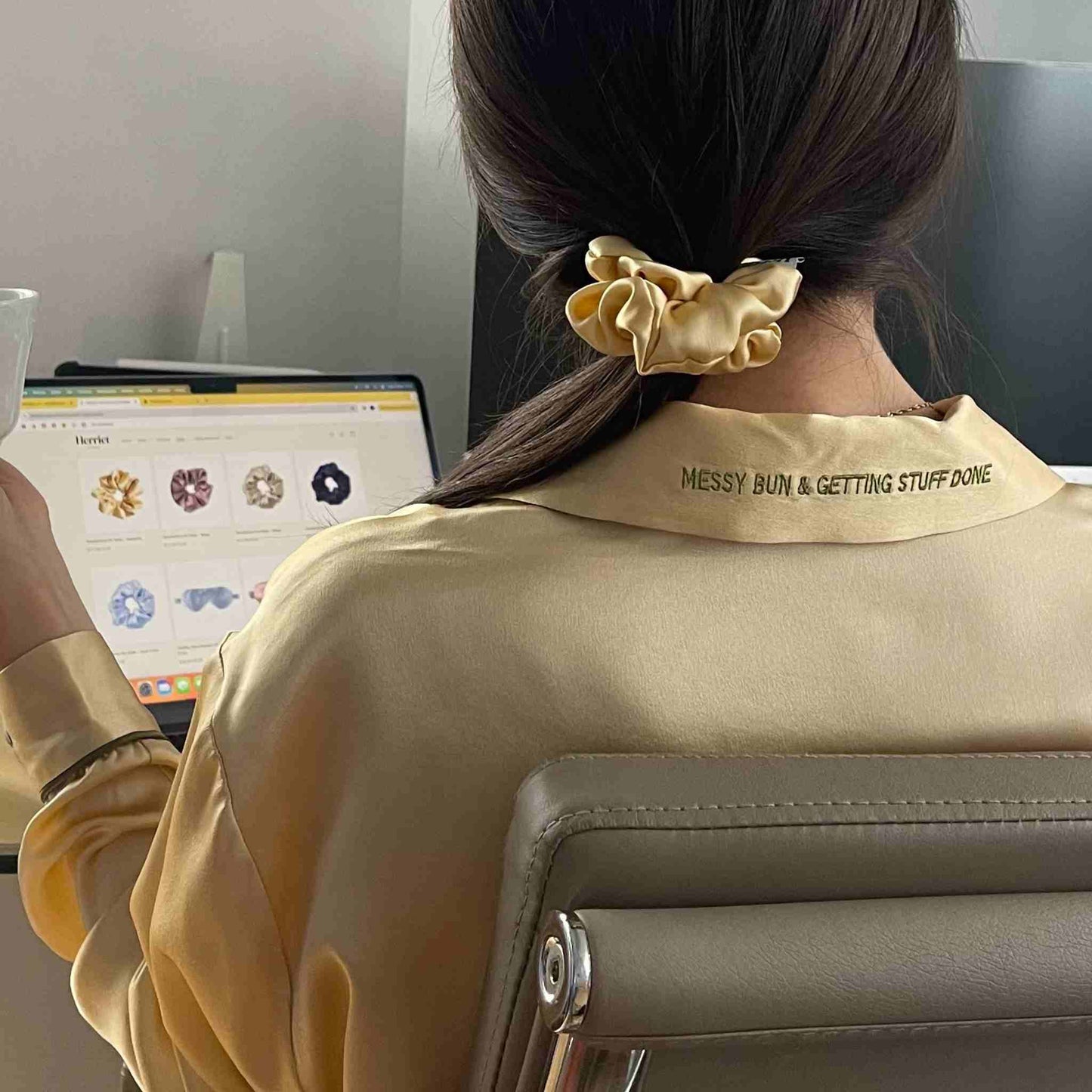 pijama de seda amarillo con frase bordada en el cuello "messy bun & getting stuff done" de herriet. La chica está de espaldas mirando el ordenador en la web de Herriet y también tiene un scrunchie de seda del mismo color del pijama